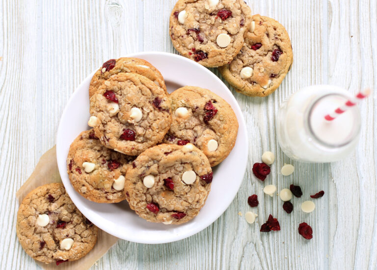 Oatmeal Cranberry cookies on plate and wooden tabel with white chocolate chips and cranberries next to glass of milk with straw